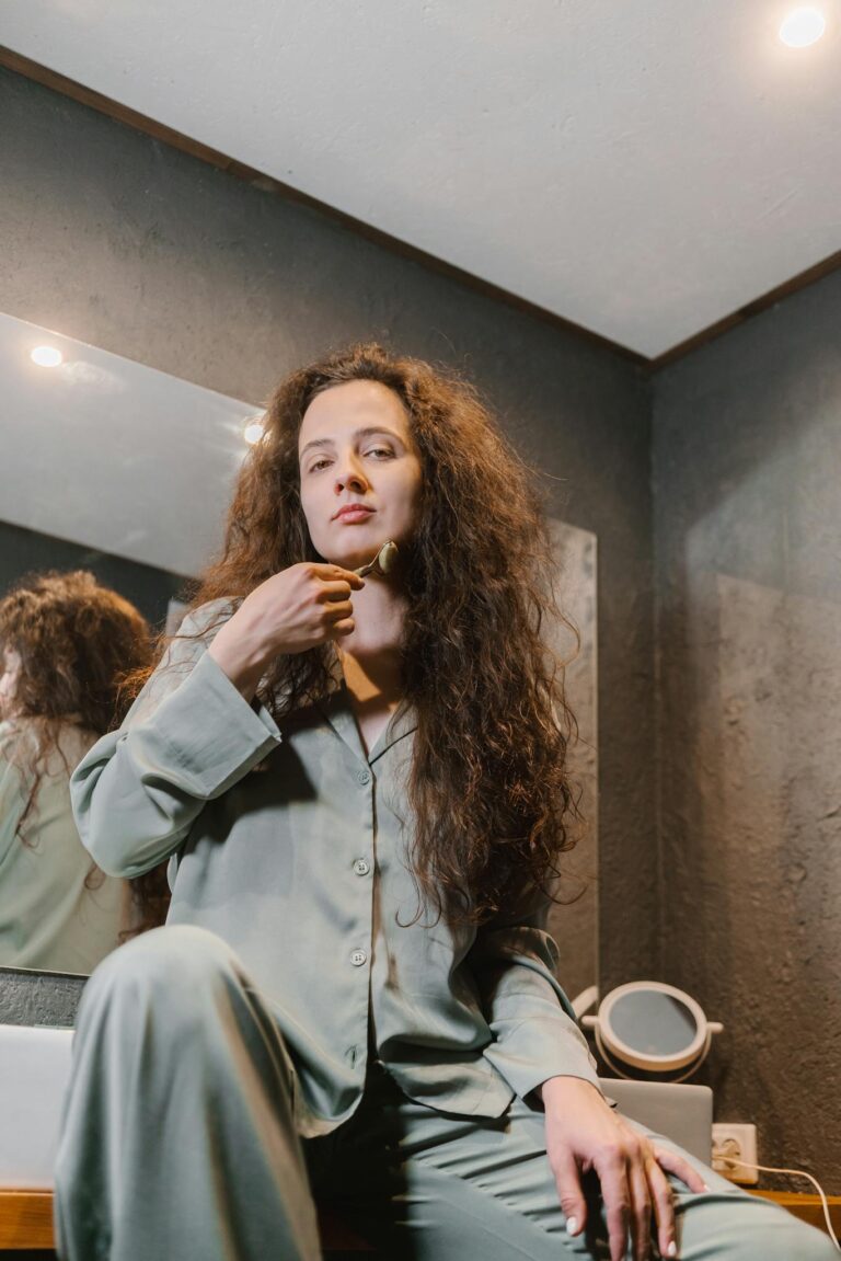 Woman in comfortable pajamas using a jade roller in a cozy, dimly lit room for a calming skincare routine.